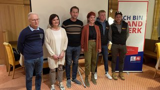 News Sechs Personen stehen vor einer Leinwand und einem Banner in einem Veranstaltungsraum