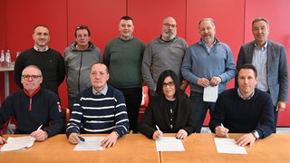 News Zehn Personen bei Vertragsunterzeichnung vor roter Wand