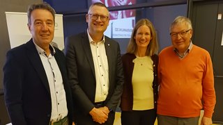 News Vier Personen in formeller Kleidung stehen vor einem Fenster und lächeln