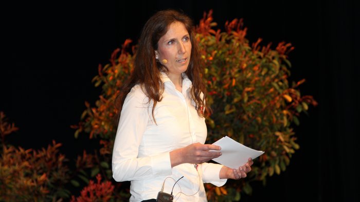 Landesversammlung des Südtiroler Handwerks Frau hält Notizen und spricht mit Mikrofon auf einer Bühne vor Pflanzen