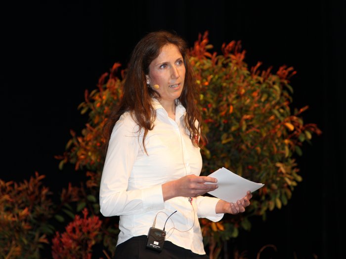 Landesversammlung des Südtiroler Handwerks Frau hält Notizen und spricht mit Mikrofon auf einer Bühne vor Pflanzen