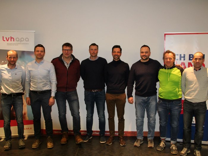 Neuer Obmann für die Berufsgemeinschaft Gruppenfoto von acht Männern vor Werbebannern in einem Veranstaltungsraum