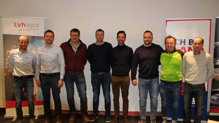 Neuer Obmann für die Berufsgemeinschaft Gruppenfoto von acht Männern vor Werbebannern in einem Veranstaltungsraum