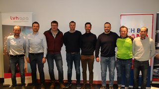 News Gruppenfoto von acht Männern vor Werbebannern in einem Veranstaltungsraum