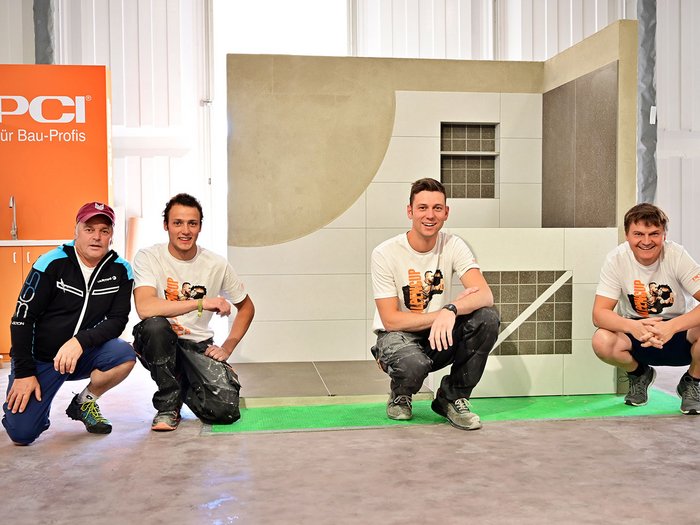 Foto 2 - Martin Domanegg und Alexander Taschler mit ihrem Team © Roland Strimmer Vier Männer knien vor einer Wand mit Fliesenmuster und PCI-Bauprofis-Schild