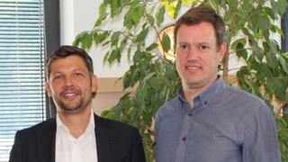 News Zwei Männer in Business-Kleidung vor grüner Pflanze und Fenster