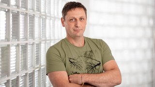 News Mann mit grüner T-Shirt steht vor eine Glaswand mit verschränkten Armen