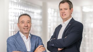 News Zwei Männer in Anzügen mit verschränkten Armen vor hellem Hintergrund