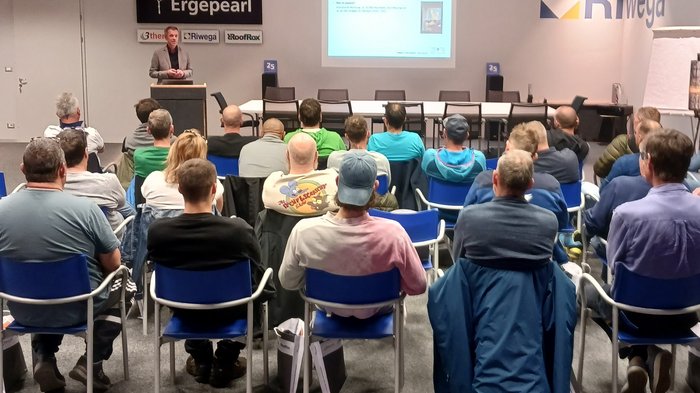 Jährlicher Pflichtkurs der Kaminkehrer/innen Person hält Vortrag vor Publikum in Konferenzraum mit Firmenlogos an der Wand