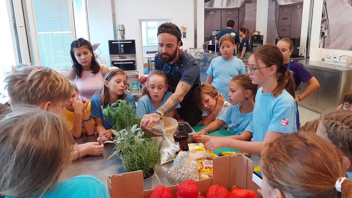 Kids-Academy 2024: Das große Finale Kinder und Mann lernen in einer Küche mit frischen Paprikaschoten und Kräutern