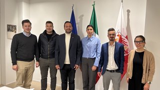 News Sechs Personen stehen nebeneinander vor drei Flaggen in einem Büro