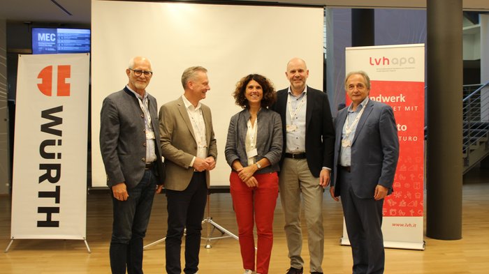 Kfz-Vierländertreffen in Bozen: Wertvoller Austausch und Dialog im Automobilsektor Fünf Personen stehen vor einer Leinwand und Werbebannern bei einer Veranstaltung