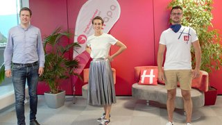 News Drei Menschen stehen vor roter Wand mit Pflanzen und Sitzgelegenheiten