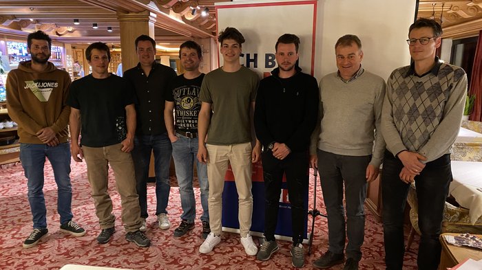 Auf jung folgt jung Acht Männer stehen in einem Raum mit rotem Teppich und Holzbalken an der Decke