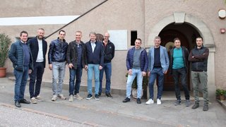 © lvh.apa Elf Männer stehen vor einem Gebäude mit Pflanze und Schild an der Wand