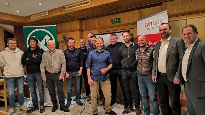 Gruppenfoto von zwölf Männern in einem Raum mit Holzverkleidung und Bannern im Hintergrund