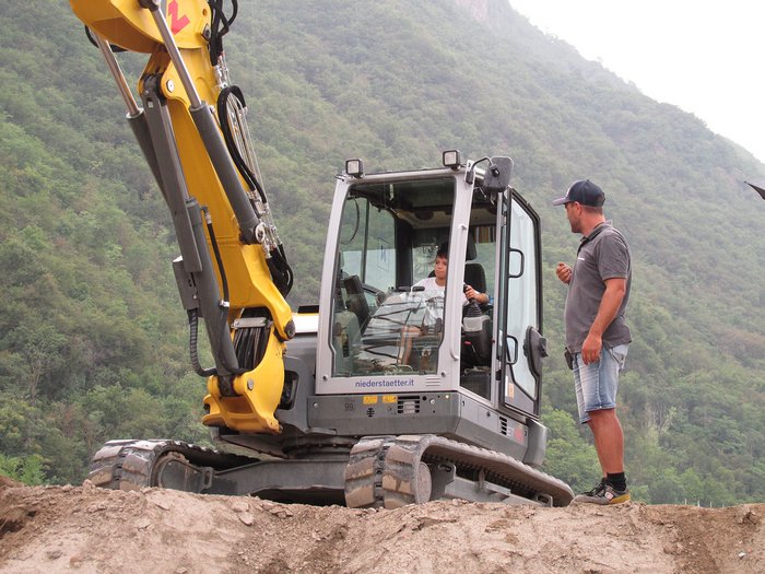 Kinder und Jugendliche lernen die Welt der Baumaschinen Kind lernt Baggerfahren mit Erwachsenem im Freien vor bewaldetem Hügel