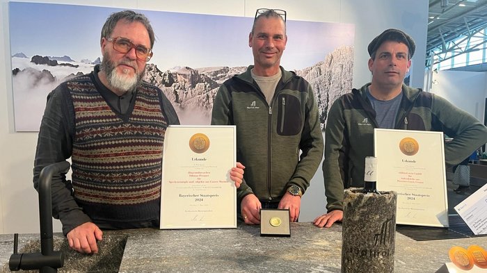 Due aziende altoatesine premiate con il “Bayrischer Staatspreis” Tre uomini con certificati e medaglia davanti a bancone in marmo con sfondo montano