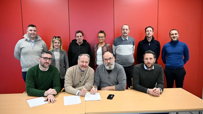 Südtiroler Bauhandwerk setzt Zeichen Gruppe von elf Personen vor roter Wand, zwei Männer unterschreiben Dokumente am Tisch