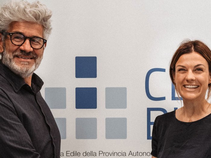 Markus Bernard an der Spitze der Bauarbeiterkasse der Autonomen Provinz Bozen Zwei lächelnde Personen vor einer Wand mit blauer Grafik und Schrift