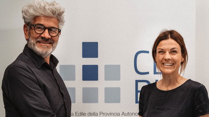 Markus Bernard an der Spitze der Bauarbeiterkasse der Autonomen Provinz Bozen Zwei lächelnde Personen vor einer Wand mit blauer Grafik und Schrift