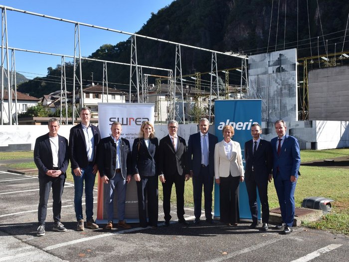 Südtiroler Wirtschaftsring und Alperia erweitern Energieangebote für SWREA-Mitgliedsunternehmen Neun Personen stehen vor einer Baustellenkonstruktion mit Bannern und bewaldeten Bergen im Hintergrund