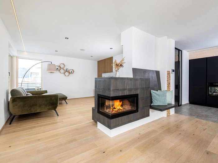 Wärme, die bleibt – Warum der Kachelofen gerade jetzt eine Renaissance erlebt Moderner Wohnraum mit Kamin, grünem Sofa und Holzboden