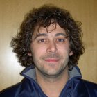 Zahntechniker Porträt eines Mannes mit lockigem Haar in blauer Jacke vor Holzwand