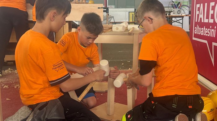 “Avventura artigianato”: l'iniziativa per i giovani talenti all'Intercable Arena di Brunico Bambini lavorano insieme su un progetto in legno con magliette arancioni