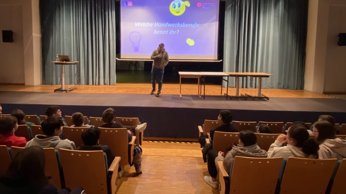 Mittelschulen Mals und Graun im Vinschgau: Handwerk & Gastgewerbe hautnah erleben. Eine Initiative der Junghandwerker/innen und der HGJ. Mann spricht vor Publikum in einem Vortrag über Handwerksberufe