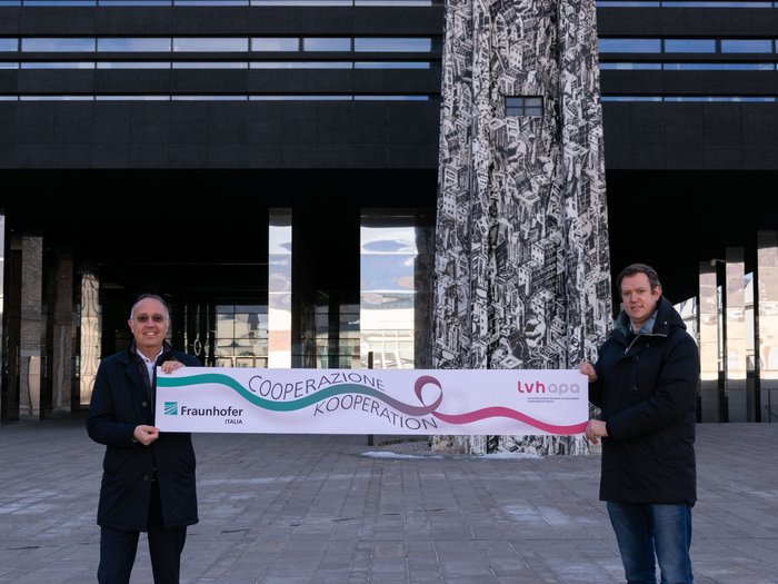 Kooperation zwischen lvh und Fraunhofer Italia verlängert Zwei Männer halten ein Banner mit den Logos von Fraunhofer Italia und LVH APA vor einem modernen Gebäude
