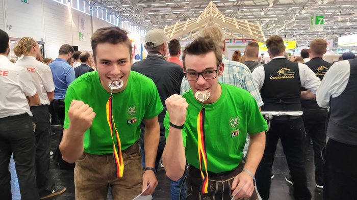 Foto 2 - Manuel Erschbaumer + Gabriel Mair © Südtiroler Zimmerer Zwei junge Männer bei einer Veranstaltung mit Medaillen im Mund und erhobener Faust