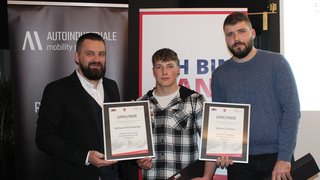 News Drei Männer halten Urkunden bei einer Auszeichnung mit Bannern im Hintergrund