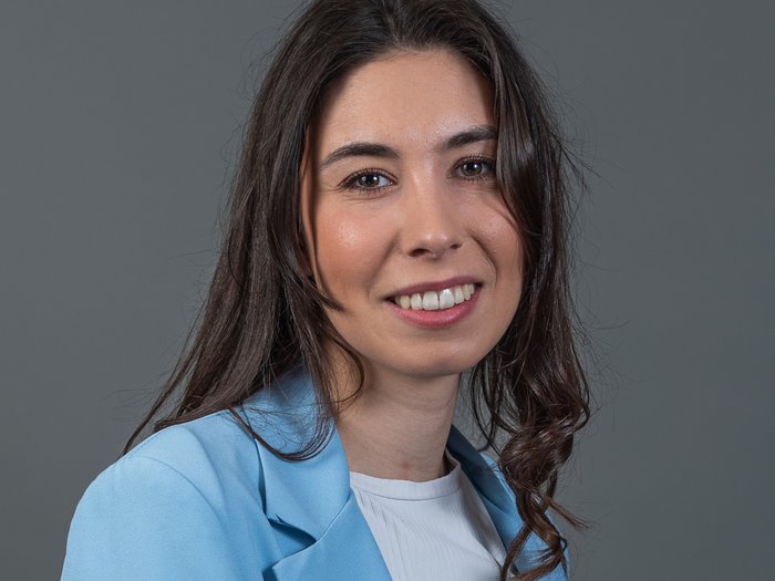 3670 neue junge Fachkräfte Porträt einer lächelnden Frau mit braunen Haaren und blauem Blazer vor grauem Hintergrund