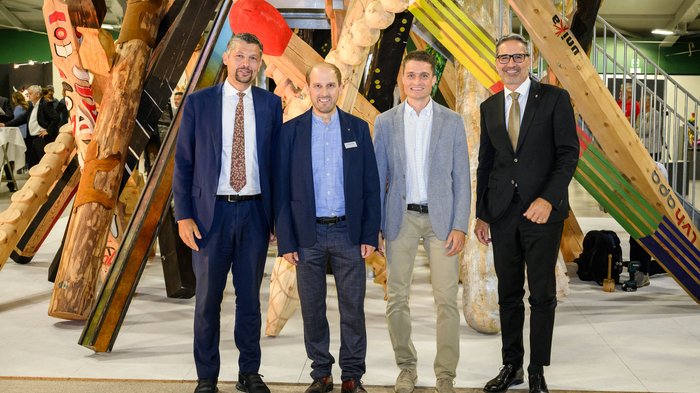 Nachbericht UNIKA Kunstmesse 2024 Vier Männer in Anzügen stehen vor einer großen Holzskulptur in Innenraum