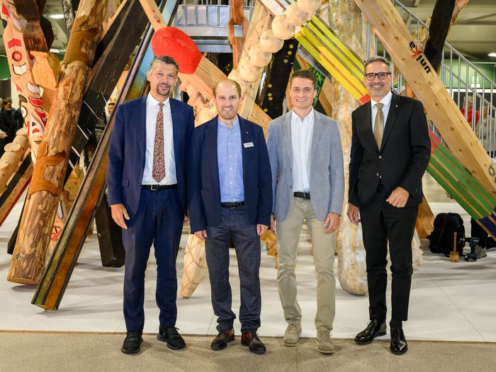 Nachbericht UNIKA Kunstmesse 2024 Vier Männer in Anzügen stehen vor einer großen Holzskulptur in Innenraum