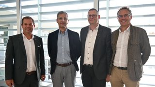 News Vier Männer in Anzügen und Hemden stehen vor großen Fenstern