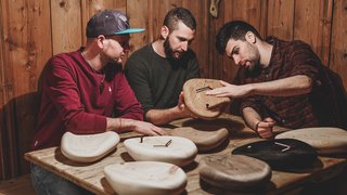 Drei Unternehmer am Tüfteln – Foto: © huamet Drei Männer besprechen handgefertigte Holzuhren an einem Holztisch