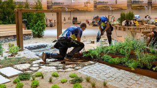 News Zwei Männer gestalten eine Gartenanlage mit Pflanzen und Holzbelag in einer Halle