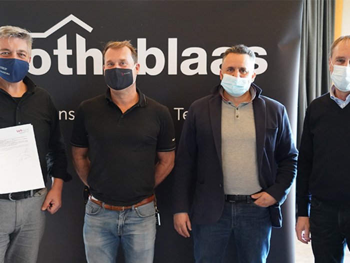 Holzbau in Südtirol: Neue Kooperation im Zeichen der Innovation Vier Männer mit Gesichtsmasken vor Firmenbanner, einer hält ein Dokument