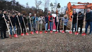 News Gruppe bei Spatenstich mit roten Schaufeln auf Baustelle mit Baggern