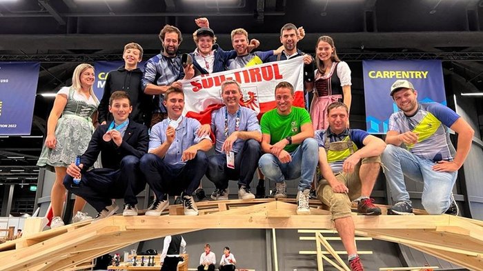 WorldSkills 2022 Special Edition: Zimmerer Marcel Bolego holt Silbermedaille Gruppenfoto von Personen auf einer Holzkonstruktion mit Südtirol-Flagge bei Zimmermannswettbewerb