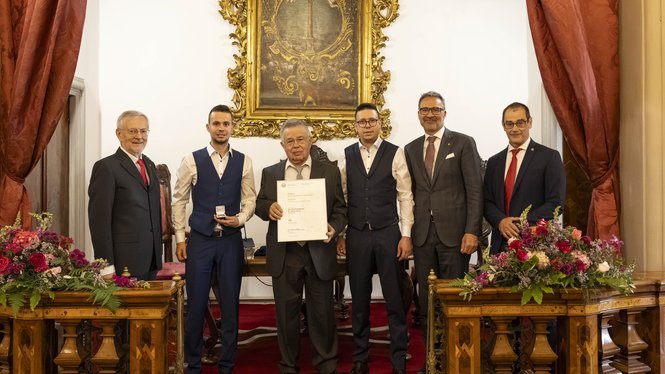 Home Sechs Männer in förmlicher Kleidung bei einer Zertifikatsübergabe vor einem prunkvollen Bild