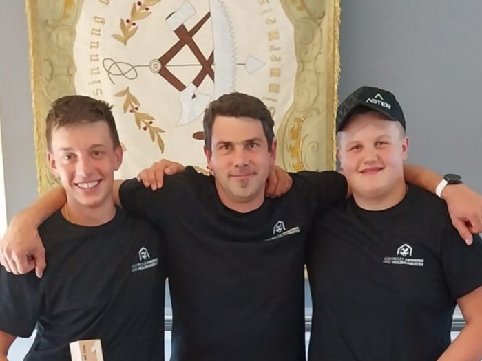 Wenn Holz Zukunft baut Drei Männer in schwarzen T-Shirts posieren vor einem Wappen an der Wand