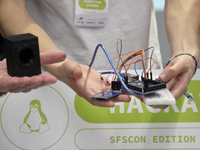 Menti creative per risolvere le sfide dell’artigianato Persona tiene circuito elettronico con fili e piccolo altoparlante davanti a poster Hackathon