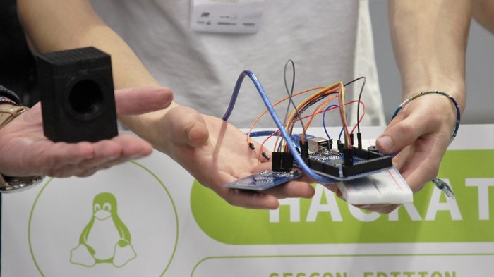 © lvh.apa Person hält elektronische Schaltung mit Kabeln und kleinem Lautsprecher vor Hackathon-Plakat