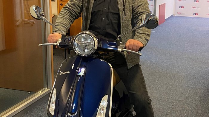 Generation-H-Vespa wird im Vinschgau unterwegs sein Mann mit Maske sitzt auf blauem Roller in Büroflur