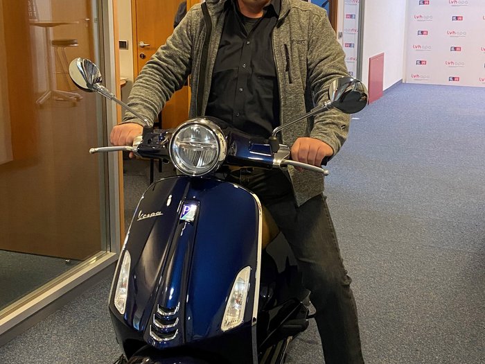 Generation-H-Vespa wird im Vinschgau unterwegs sein Mann mit Maske sitzt auf blauem Roller in Büroflur