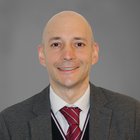 Öffentliche Aufträge Porträt eines lächelnden Mannes in Anzug und roter Krawatte vor grauem Hintergrund