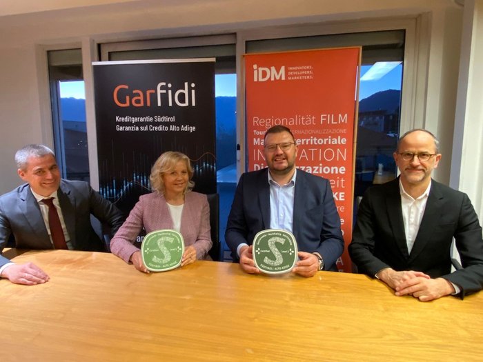L’impegno verso la sostenibilità viene premiato: Garanzie di prestito agevolate per aziende con il Marchio Sostenibilità Alto Adige Quattro persone sedute al tavolo con certificati davanti a banner Garfidi e IDM